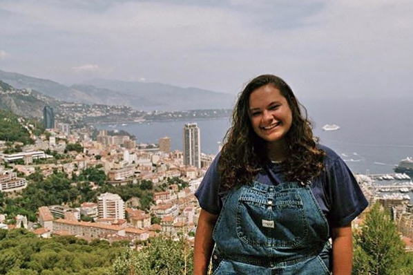 Sophie Moser smiles while wearing overalls in front of a city and ocean landscape